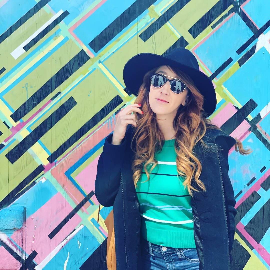 A beautiful young woman in a hat and sunglasses wearing a green shirt stands in front of a colorful background